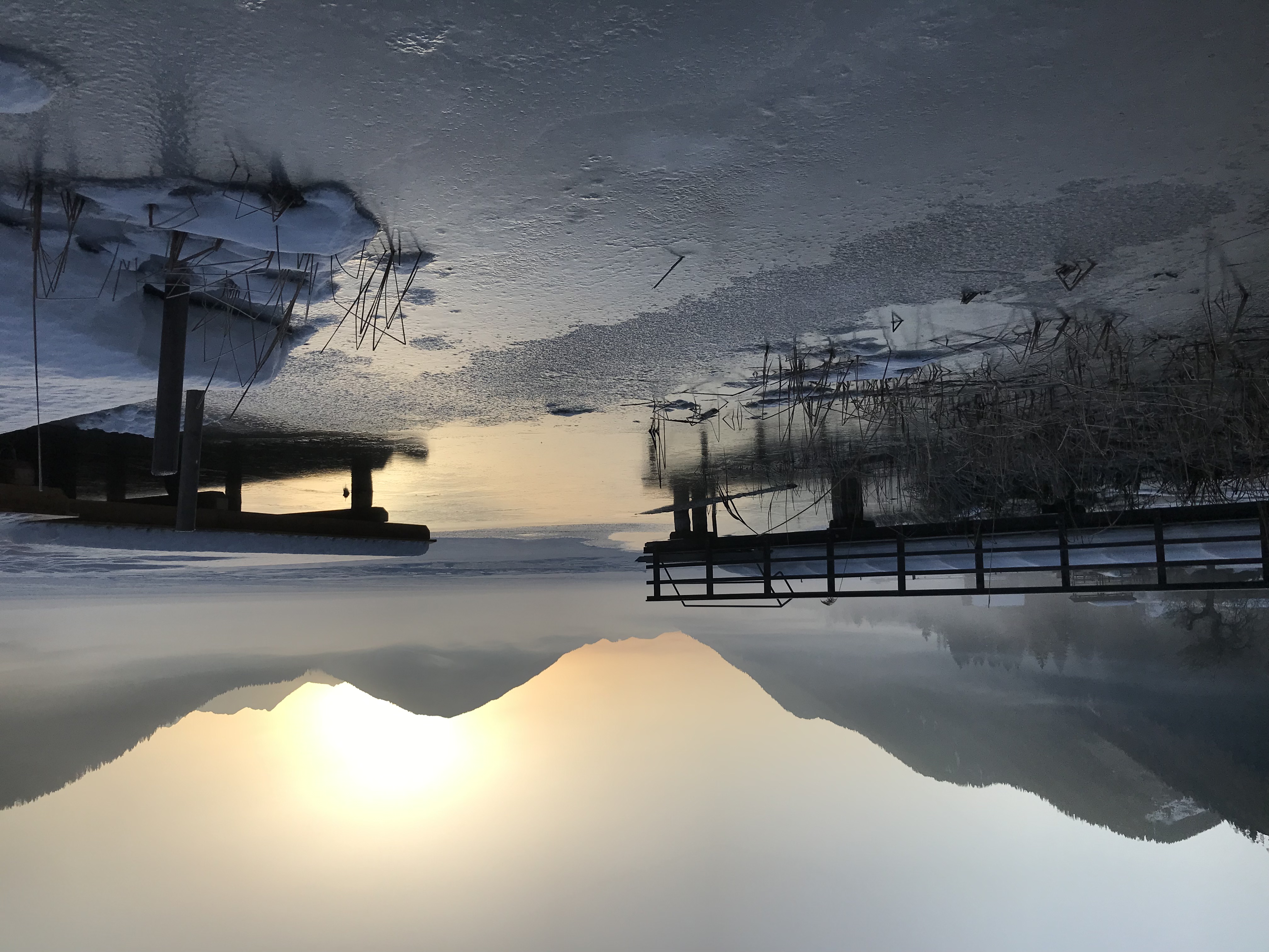 Teilweise zugefrorenes Wasser, zwei verschneite Stege und Nebel unterhalb der über den Bergen aufgehenden Sonne.