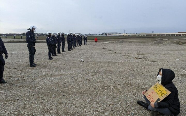 Polizeiabsperrung, im Bild rechts davon sitzt ein Mensch mit Maske und Schild: Öffiliebe statt Klimakrise