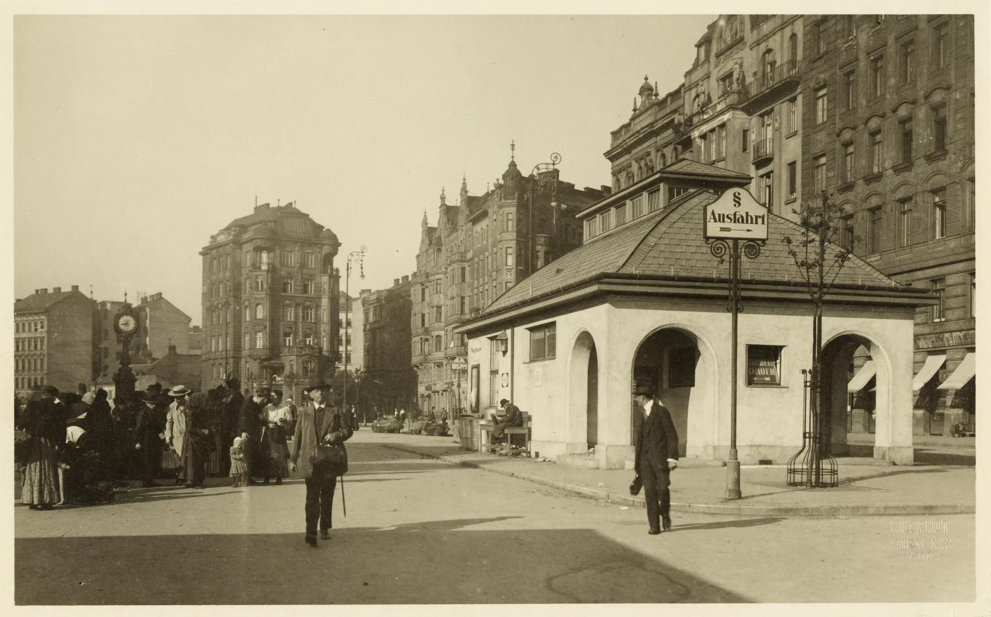 Schwarzweißfoto eines befestigten Platzes in der Stadt. Mann mit Hut und Spazierstock, dahinter eine Menschenmenge. Rechts ein kleines Gebäude mit „Männer“ und „Frauen“ Beschriftung – also ein WC. Im Hintergrund mehrstöckige Gründerzeithäuser.