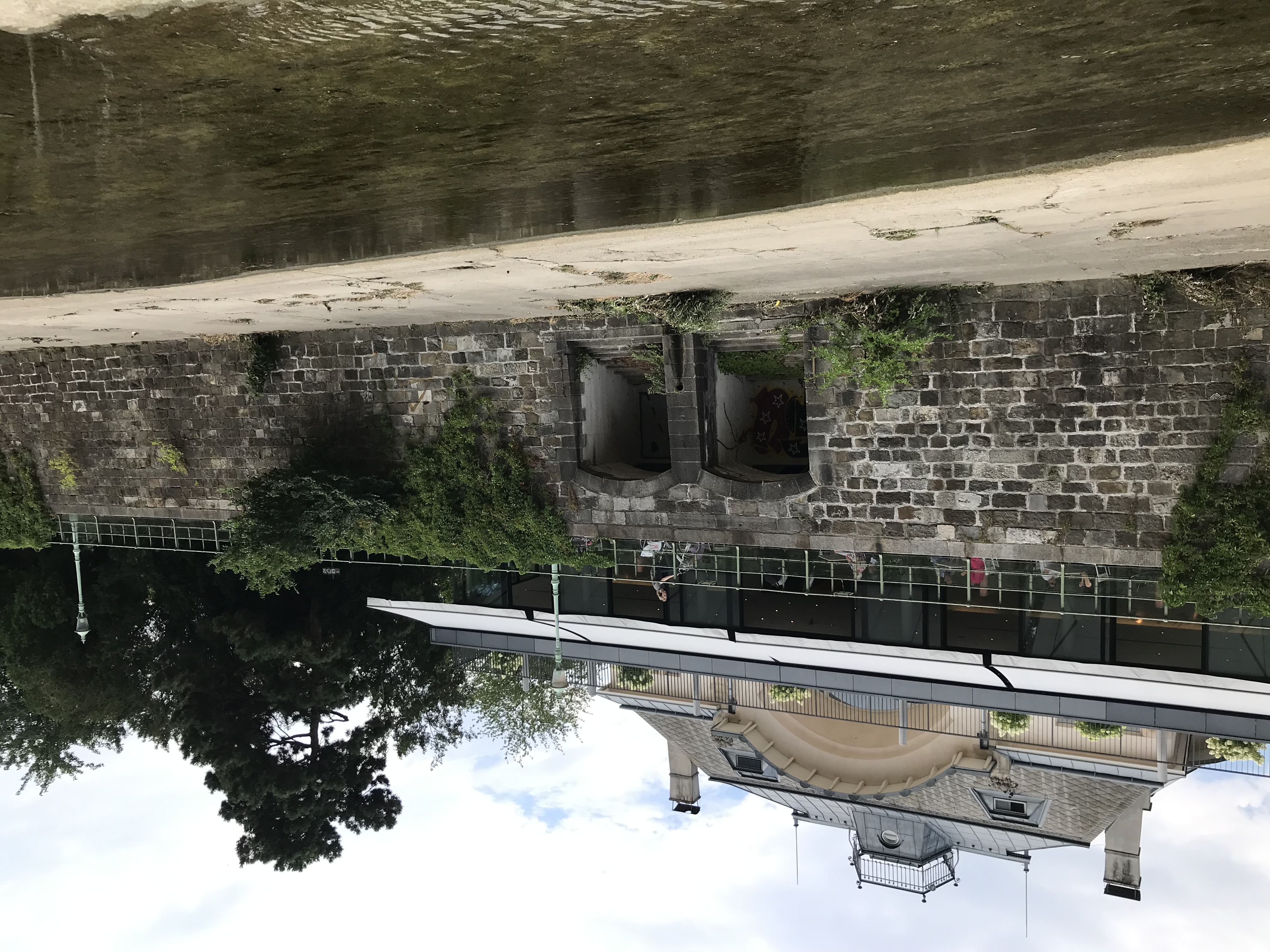 Senkrechte Wand aus Steinquadern, die ein Flussbett begrenzen. In der Wand zwei Torbögen mit Stufen. Oberhalb ist ein älteres, repräsentatives aber eher kleines Gebäude zu sehen.