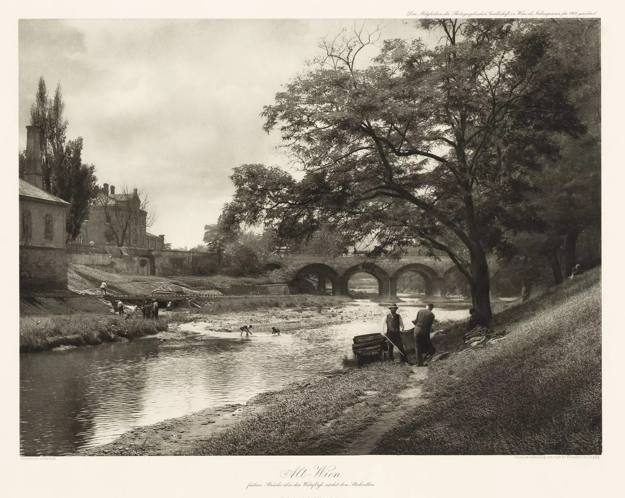 Schwarzweißfoto: Ein von grasigen Böschungen begrenztes Flussbett. Rechts unter einem Laubbaum zwei Männer mit Hüten, welche einen Handwagen beladen. Im Hintergrund eine Bogenbrücke aus Steinquadern. Am linken Ufer ein Gebäude im Anschnitt und im seichten Flussbett spielende Kinder.