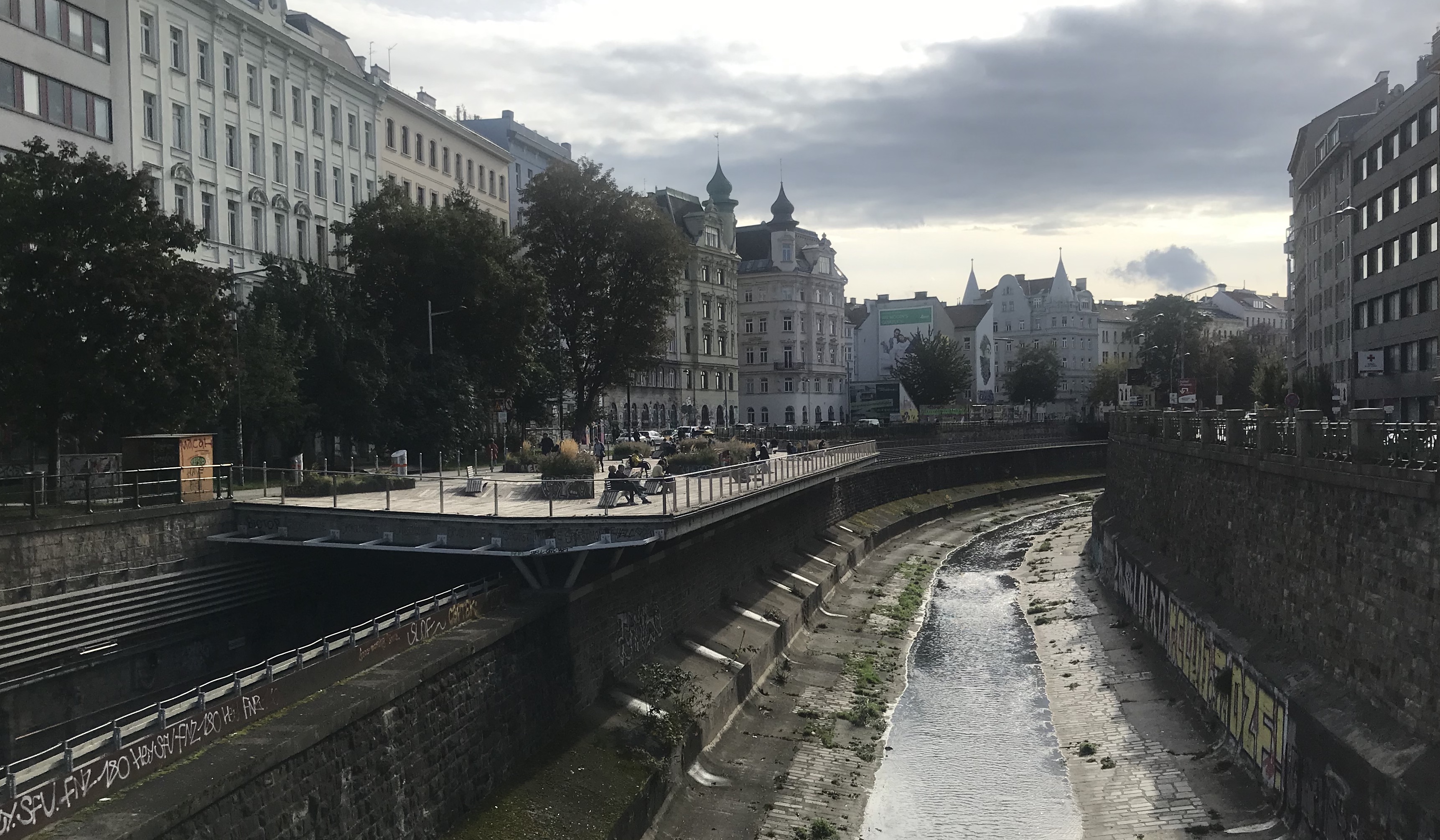Blick in den Wienflusskanal, links eine Terrasse mit Menschen im Nachmittagslicht.
