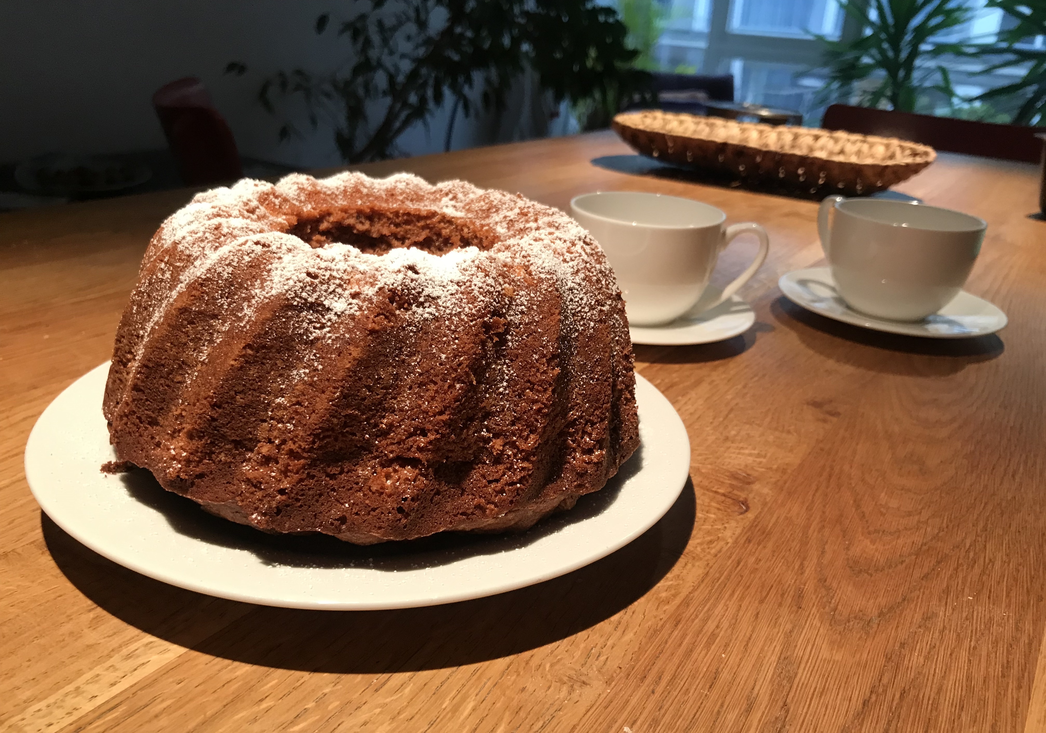 Überzuckerter Gugelhupf auf einer Holzplatte. Kaffeetassen im Hintergrund.