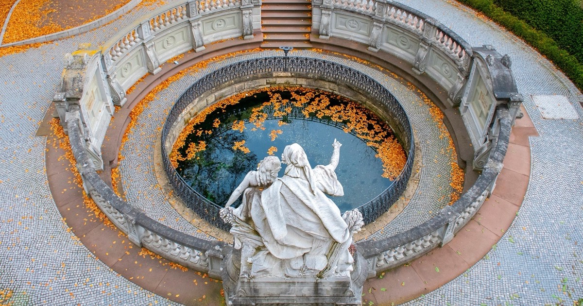 Farbfoto: Donauquelle in Donaueschingen von oben, ein rundes Brunnenbecken mit Herbstlaub auf dem Wasser, umgeben von einer Balustrade, davor eine Figurengruppe aus Marmor